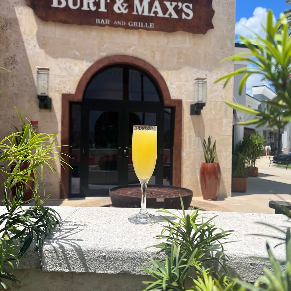 A yellow cocktail sits before the tropical, palm-lined entrance of Burt & Max’s Bar and Grille.