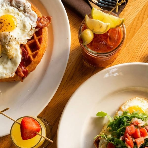 Waffle with bacon and fried eggs, avocado toast, a mimosa, and a Bloody Mary on a wooden table.