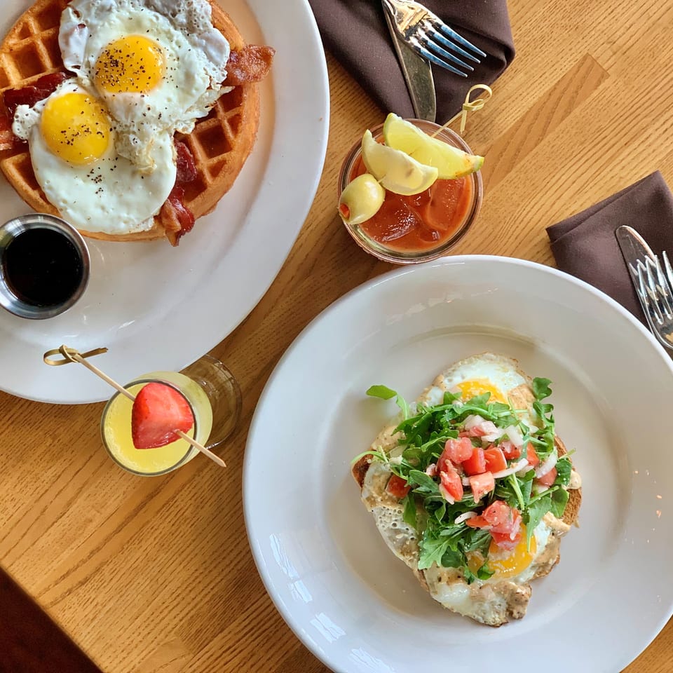 Brunch spread featuring waffles with bacon and fried eggs, an avocado toast topped with greens and tomatoes, sauces, and fruit drinks.