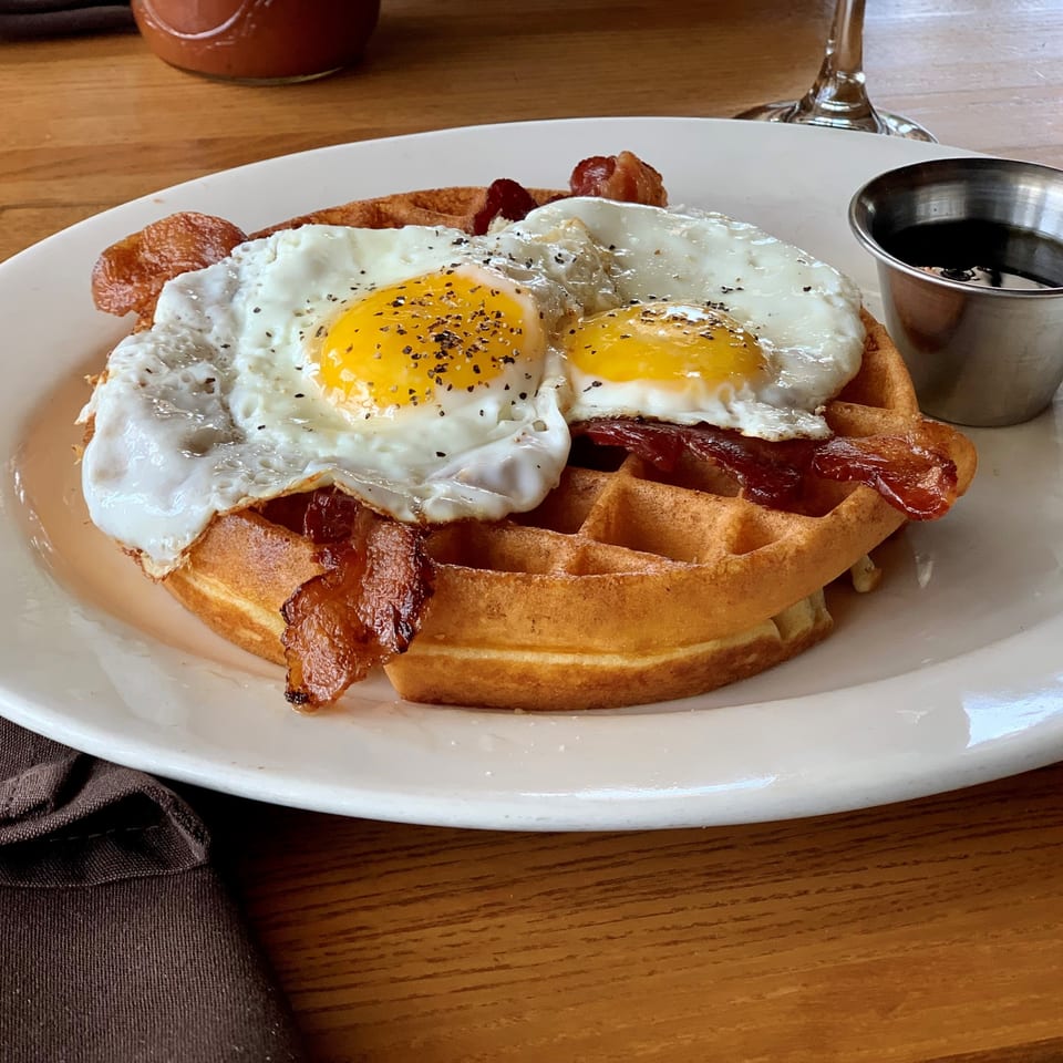 Belgian waffle topped with crispy bacon and two sunny-side-up eggs, served with maple syrup on a white plate.