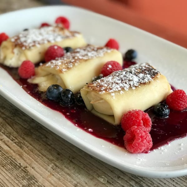 Three crepes filled with cream, drizzled with berry sauce, and garnished with fresh raspberries and blueberries.