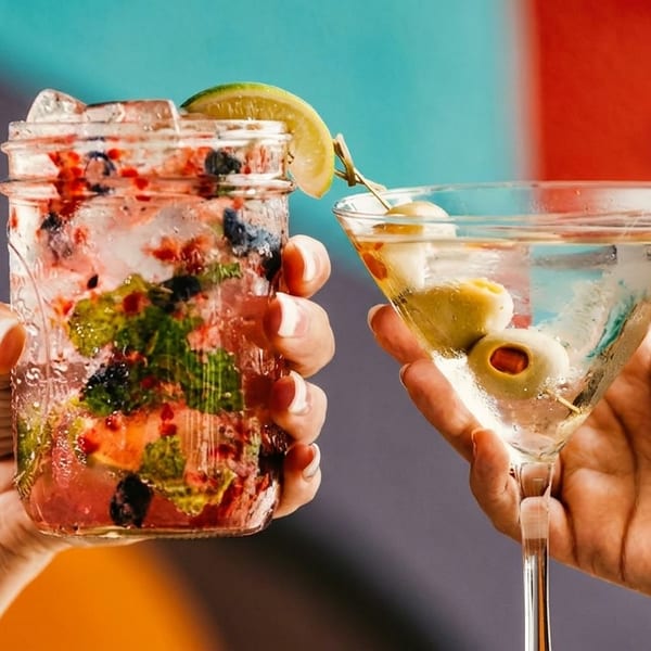 Two hands holding a berry mojito in a mason jar and a martini with olives.