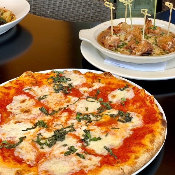 A Margherita pizza with basil and a bowl of fried chicken bites in sauce on a black table.